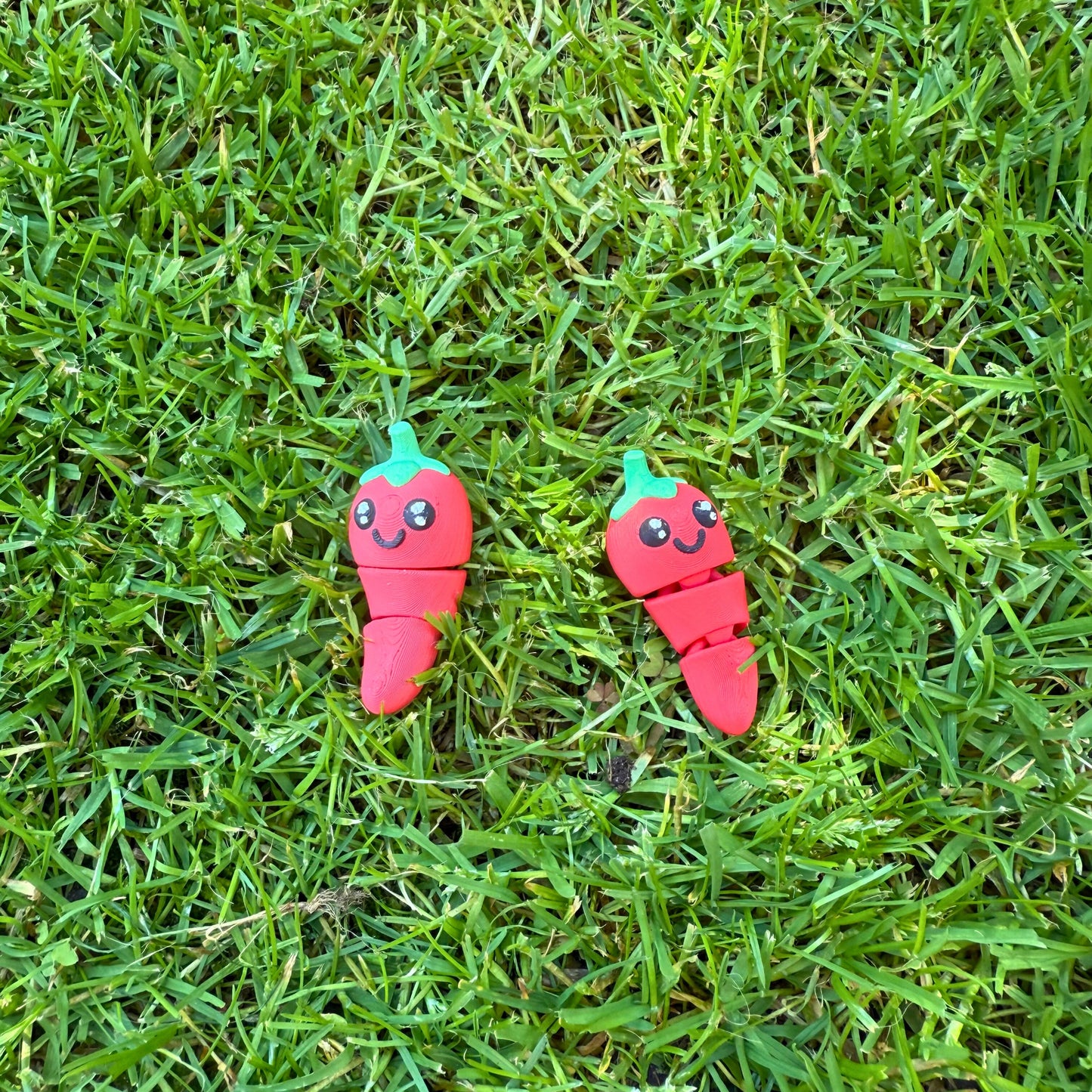 Twee 3D geprinte speelgoed chilipepers liggend in het gras, vrolijk en kleurrijk.