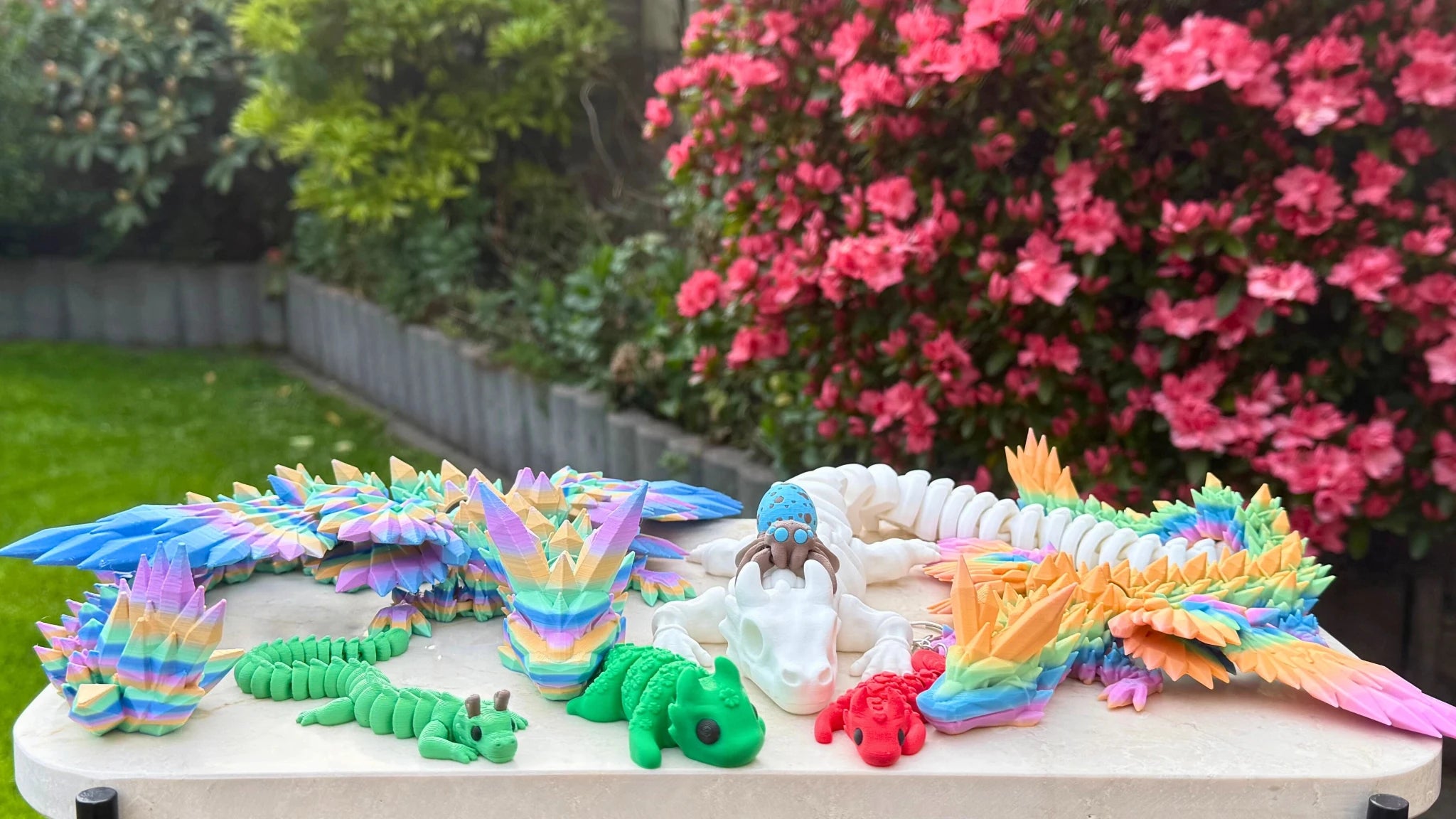 Een kleurrijke collectie 3D-geprinte speelgoedfiguren, waaronder regenboogdraken, een groene draak met horens, kleine hagedissen en een witte draak, gepresenteerd op een tafel in een bloeiende tuin.