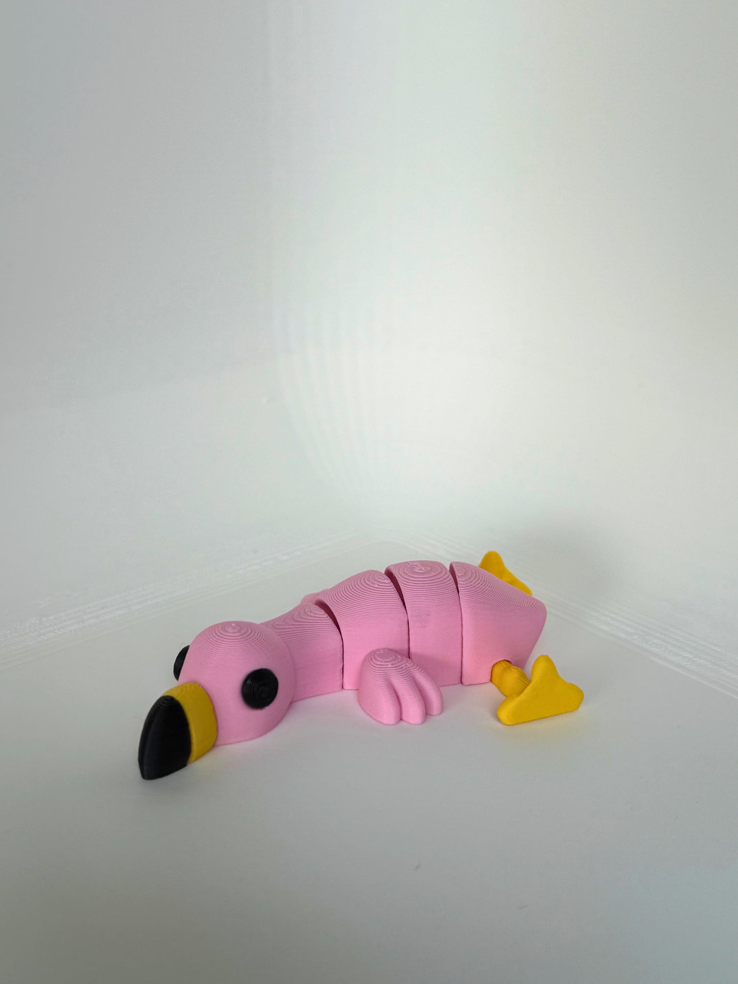Lichtroze 3D-geprinte flamingo met gele poten en zwarte snavel, liggend op een witte ondergrond.