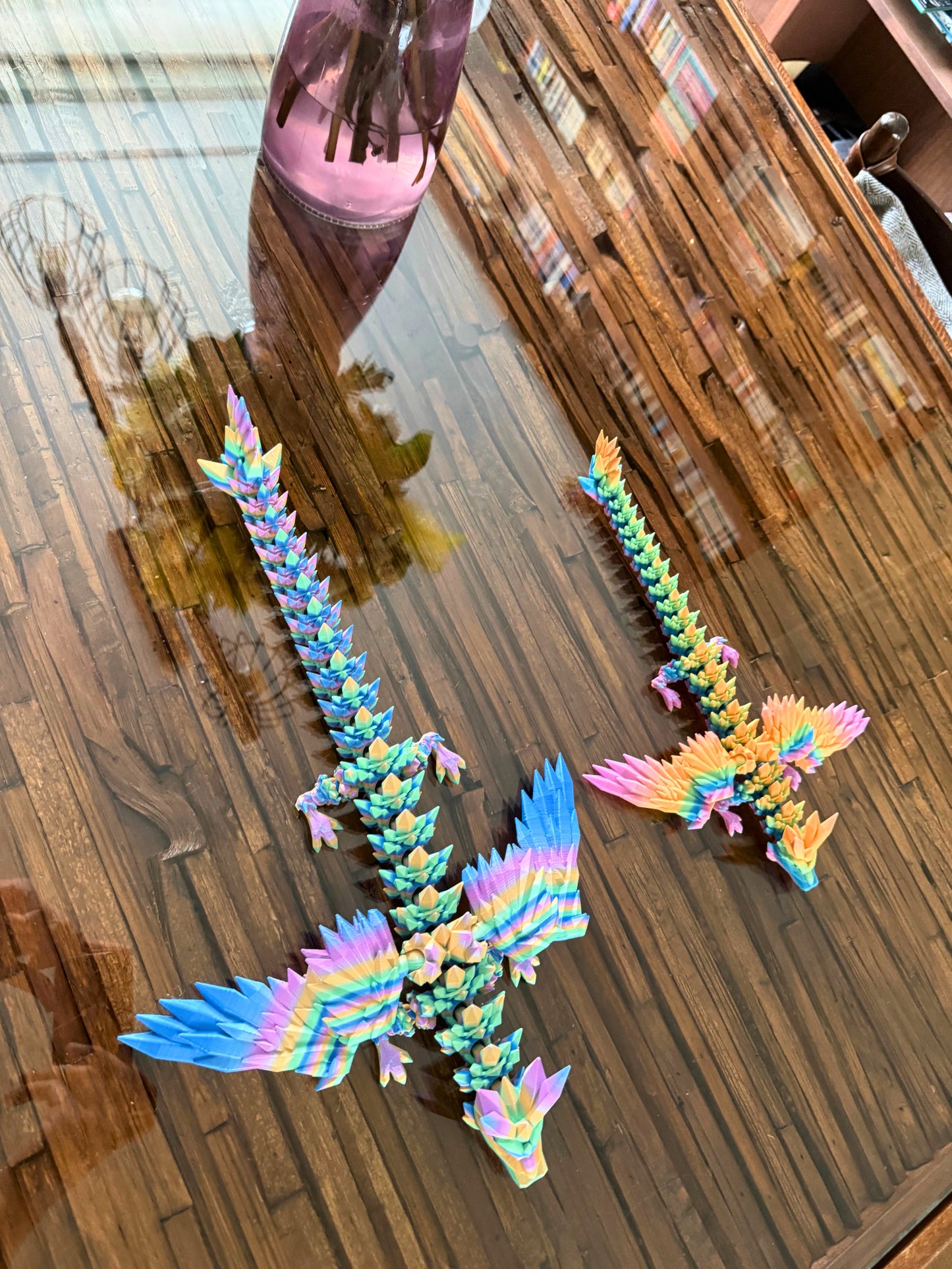 Grote articulerende regenboogdraak en kleinere versie ernaast op houten tafel, gefotografeerd van boven.