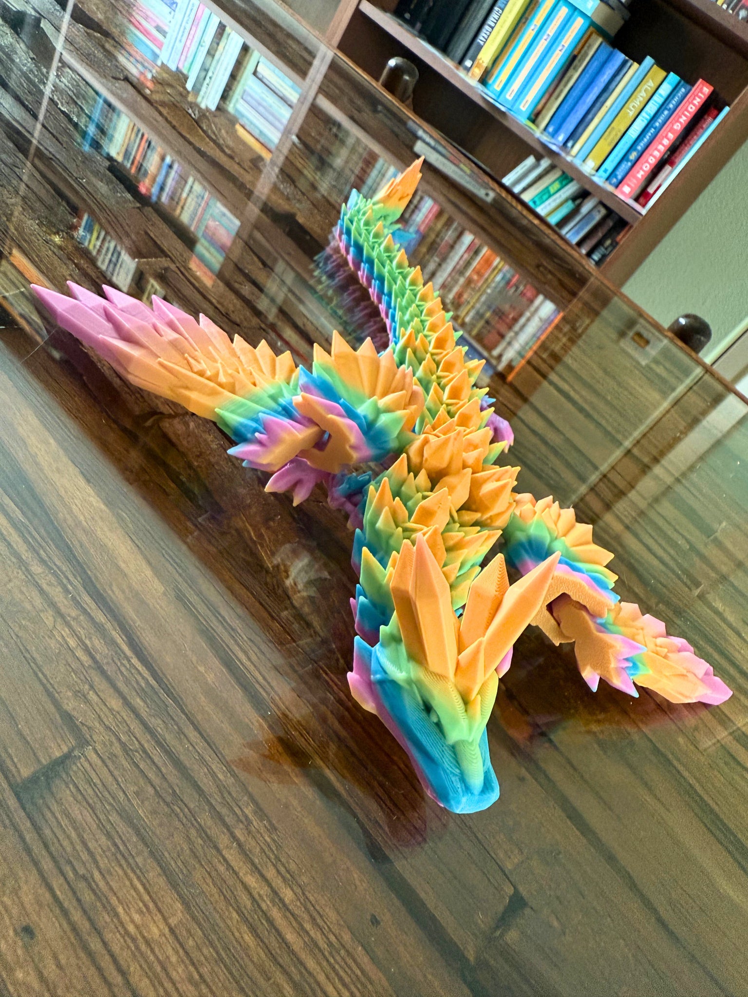 Regenboogdraak met oranje, roze en blauwe vleugels kijkt recht in de camera, liggend op een reflecterende tafel.