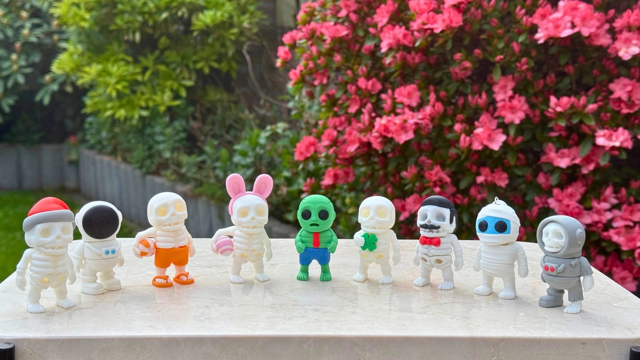 Een rij van kleurrijke 3D-geprinte skeletfiguren met verschillende thema’s, waaronder kerst, ruimte, zomer, Pasen, zombie, Sint Patrick, gentleman, mummy en astronaut, uitgestald op een tafel in een bloeiende tuin.