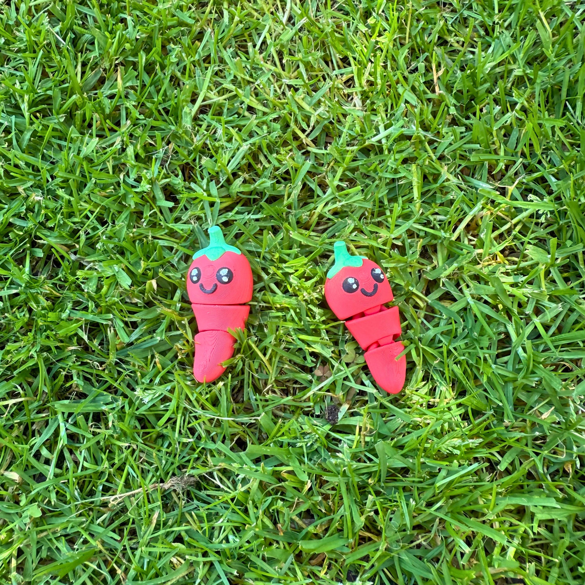 Twee 3D geprinte speelgoed chilipepers liggend in het gras, vrolijk en kleurrijk.
