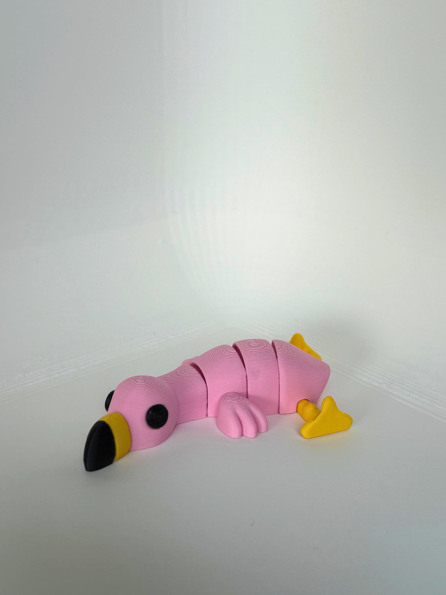 Lichtroze 3D-geprinte flamingo met gele poten en zwarte snavel, liggend op een witte ondergrond.