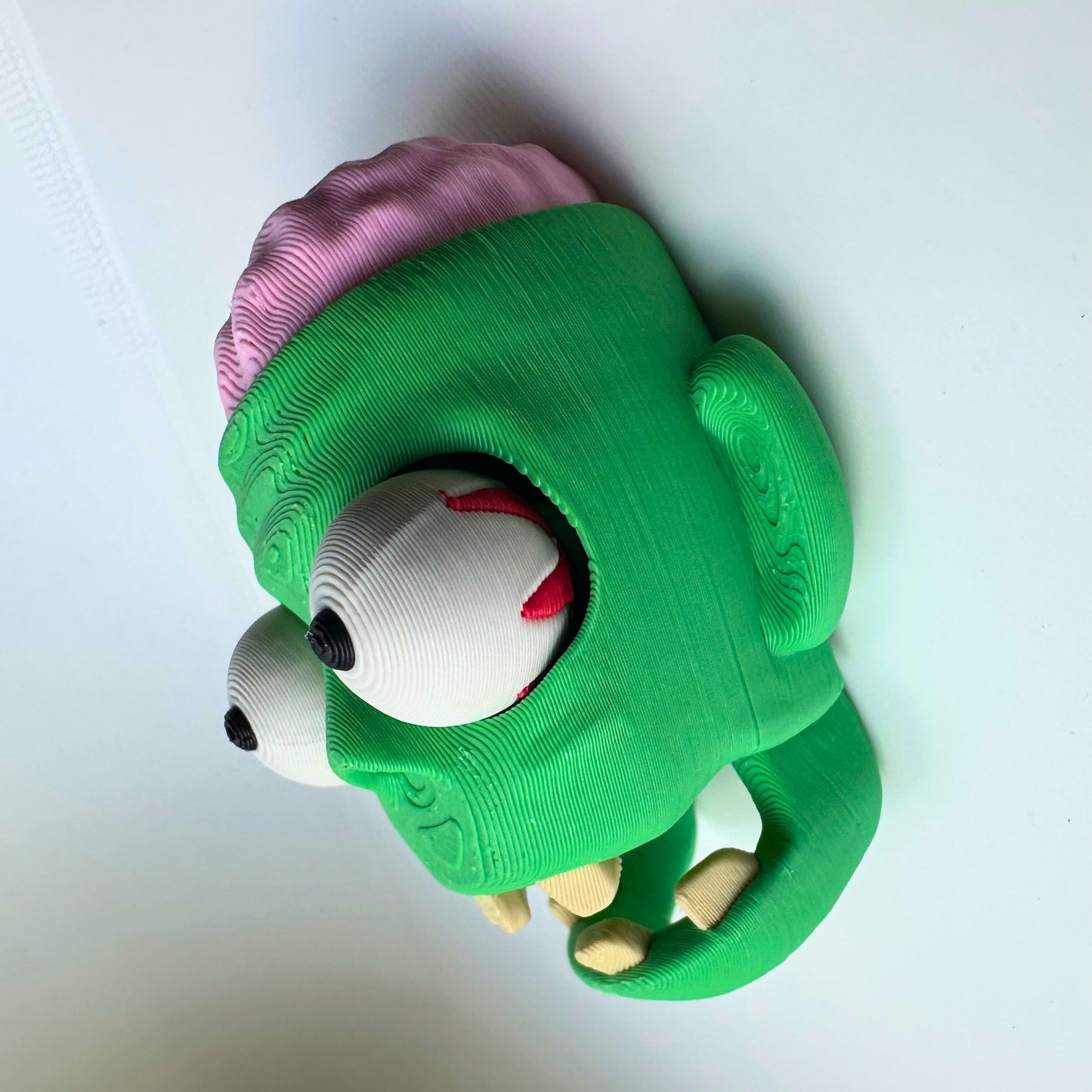 Grappige 3D-geprinte zombie kop met groene huid, grote uitpuilende ogen en roze hersenen zichtbaar bovenop, gemaakt van duurzaam PLA-plastic