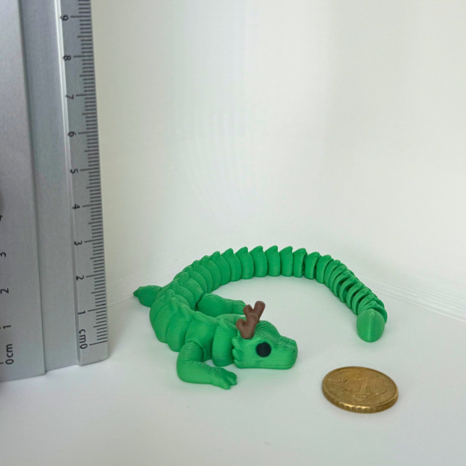 Groene 3D-geprinte draak met hoorns, liggend in een gekrulde houding naast een munt en een liniaal.