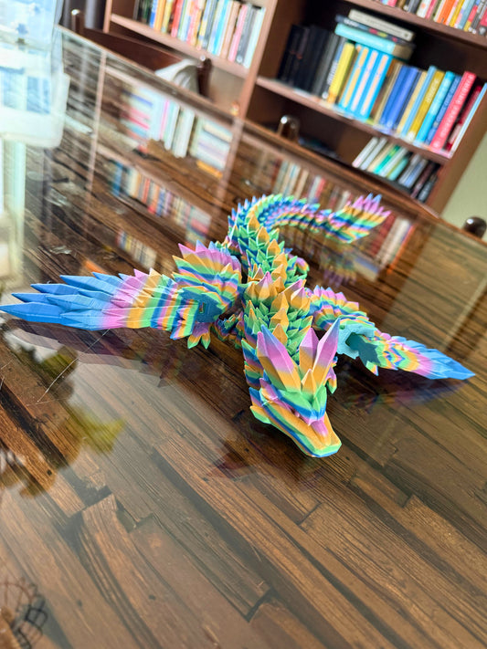Kleine articulerende regenboogdraak op een houten tafel met boekenkast op de achtergrond.